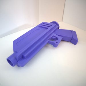 May include: A purple toy gun with a rectangular body and a handle. The toy gun has a cylindrical barrel and a trigger. The toy gun is made of a solid material and is sitting on a white surface.