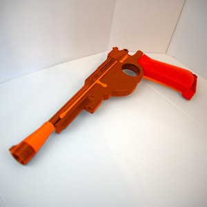May include: A toy gun in shades of brown and orange. The main body is brown, with a bright orange handle and barrel tip. The toy is made of a solid material and is designed to resemble a classic firearm.