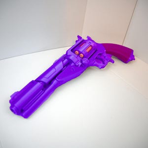 Puede incluir: Un revólver de juguete morado con un mango magenta descansa sobre una superficie blanca. La pistola de juguete presenta un diseño detallado con detalles en naranja en el cilindro y el área del gatillo. La estética general es un diseño vibrante y lúdico.