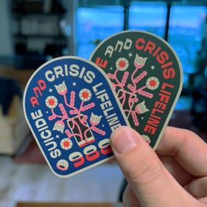 Pu&ograve; includere: Due adesivi con sfondo blu e verde. Entrambi gli adesivi presentano un telefono stilizzato con un design floreale e il testo "Suicide and Crisis Lifeline 988".