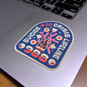 Pu&ograve; includere: Un adesivo blu e giallo con un telefono stilizzato e fiori. Il testo recita "Suicide and Crisis Lifeline 988".