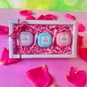 Puede incluir: Un juego de regalo de tres bálsamos labiales en una caja blanca con papel de seda rosa y una cinta roja. Los bálsamos labiales son de color lila claro, azul y rosa, cada uno con el texto "LIP FUEL". Pétalos de rosa rosas esparcidos.