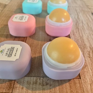 May include: Lip Fuel lip treatment in various pastel colors. The image shows several open and closed containers of lip balm. The lip balm is a light yellow color. The containers are pink, blue, and purple.