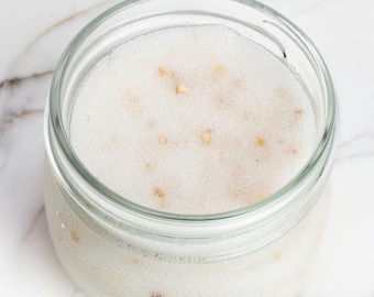 May include: A clear glass jar filled with a white, granular substance that has a textured, frosted appearance. The contents include small, scattered tan-colored particles throughout the white mixture, visible through the transparent glass walls.