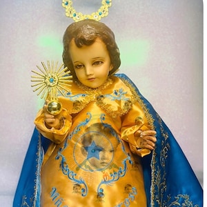 May include: A figurine of a young child dressed in a gold and blue robe with a halo above its head. The robe has gold embroidery and the text "Niño Milagroso" is visible on the front. The child is holding a golden orb in its right hand.