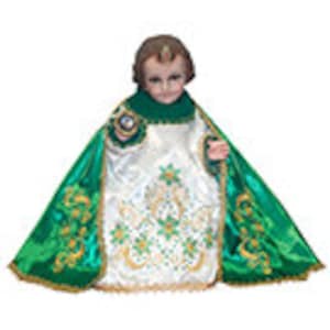 May include: A small, white figurine of a child wearing a green and gold robe. The robe is decorated with a pattern of shamrocks and has a gold trim. The figurine is standing on a white base.