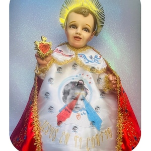 May include: A statue of a young child dressed in a white robe with a red and blue heart design. The child is wearing a gold crown and holding a gold heart. The statue is labeled "Divina Misericordia" which translates to "Divine Mercy".