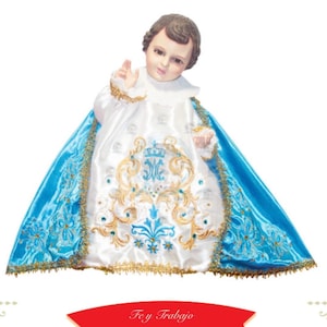 May include: A detailed figurine of a child with brown hair, wearing a white and blue satin outfit. The outfit is adorned with gold embroidery and a blue cape. The figurine has a serene expression and is holding up one hand. The text "Fe y Trabajo" is on a red banner.