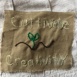 May include: A brown fabric square with the words "Cultivate Creativity" embroidered in white thread. A green plant with a brown stem is embroidered in the centre of the square.