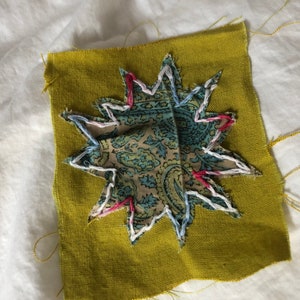 May include: A green fabric square with a star-shaped patch sewn on top. The star is made of a patterned fabric with blue, green, and brown colours. The star is outlined with white, pink, and red stitching.