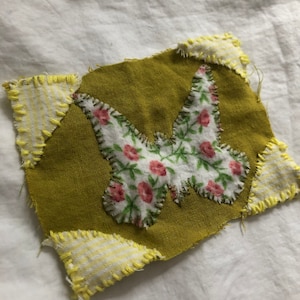 May include: A green fabric patch with a white butterfly design featuring pink flowers. The patch is surrounded by yellow and white striped fabric.