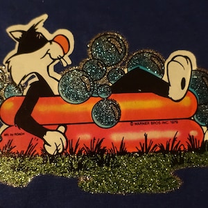 Puede incluir: Un gato de dibujos animados blanco y negro con una camisa azul se relaja en una balsa inflable roja y naranja rodeada de burbujas de purpurina azules y verdes. El texto "Warner Bros. Inc. 1979" está impreso en la balsa.