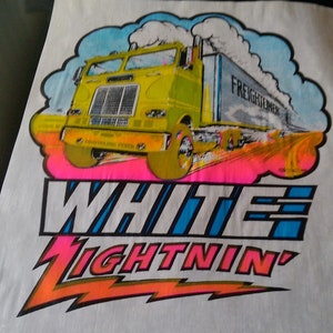 Puede incluir: Una camiseta blanca con un camión amarillo de semi-remolque conduciendo por una carretera con un brillo rosa y naranja. El camión tiene las palabras "FREIGHTLINER" en el lateral. El texto "WHITE LIGHTNIN'" está debajo del camión con un rayo.