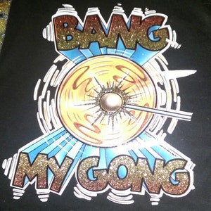 Op de afbeelding: Een zwart T-shirt met een gouden en rood glitterontwerp met de tekst "Bang My Gong". Het ontwerp toont een gong die wordt geraakt door een stok, omringd door een blauw en wit ontwerp.