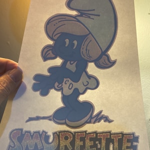 May include: A blue Smurfette cartoon character with a white dress and a blue hat. The word "SMURFETTE" is written in rainbow colors below the character.