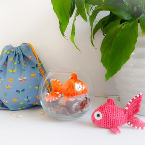 Pode incluir: Dois peixinhos de crochê, um laranja e outro rosa, com detalhes brancos. O peixinho laranja está em uma tigela de vidro transparente com elementos decorativos marrons. O peixinho rosa está sobre uma superfície branca. A imagem também inclui uma bolsa de cordão azul com um padrão de libélula.