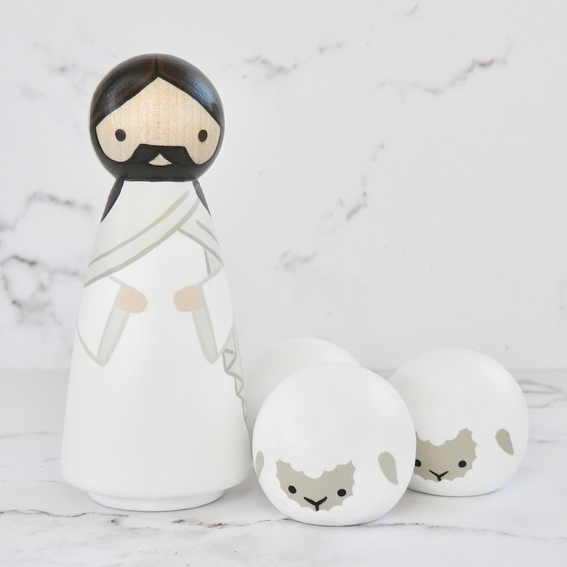 May include: A wooden doll depicting Jesus in a white robe with a gray sash, standing with two white wooden sheep with gray faces.