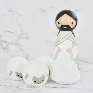May include: A wooden figurine of Jesus wearing a white robe with a brown sash, standing with three white wooden sheep figurines.