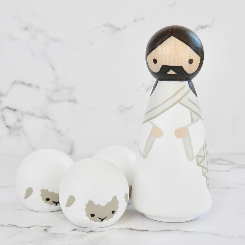 May include: A wooden figurine of Jesus wearing a white robe with a brown sash, standing with three white wooden sheep figurines.
