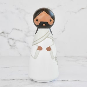 May include: A wooden peg doll painted to resemble a person wearing a white robe with a brown sash. The doll has a brown face, black hair, and a black beard and mustache.