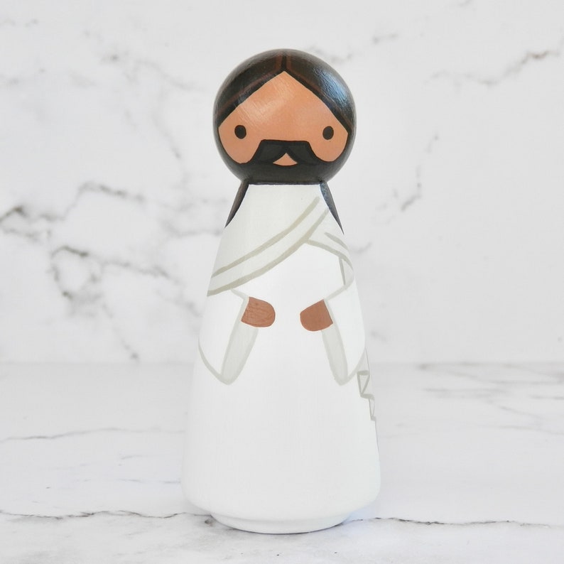 May include: A wooden peg doll painted to resemble a person wearing a white robe with a brown sash. The doll has a brown face, black hair, and a black beard and mustache.