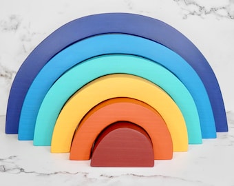 May include: A rainbow stacking toy made of wood. The toy is made up of six arches in different colors: blue, light blue, teal, yellow, orange, and brown. The arches are stacked on top of each other, with the largest arch at the bottom and the smallest arch at the top.