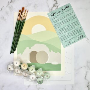 May include: A paint-by-numbers kit with a landscape design, including a canvas, paint pots, and brushes. The canvas features a yellow sun, green hills, and a tomb. A card with painting tips is also included.