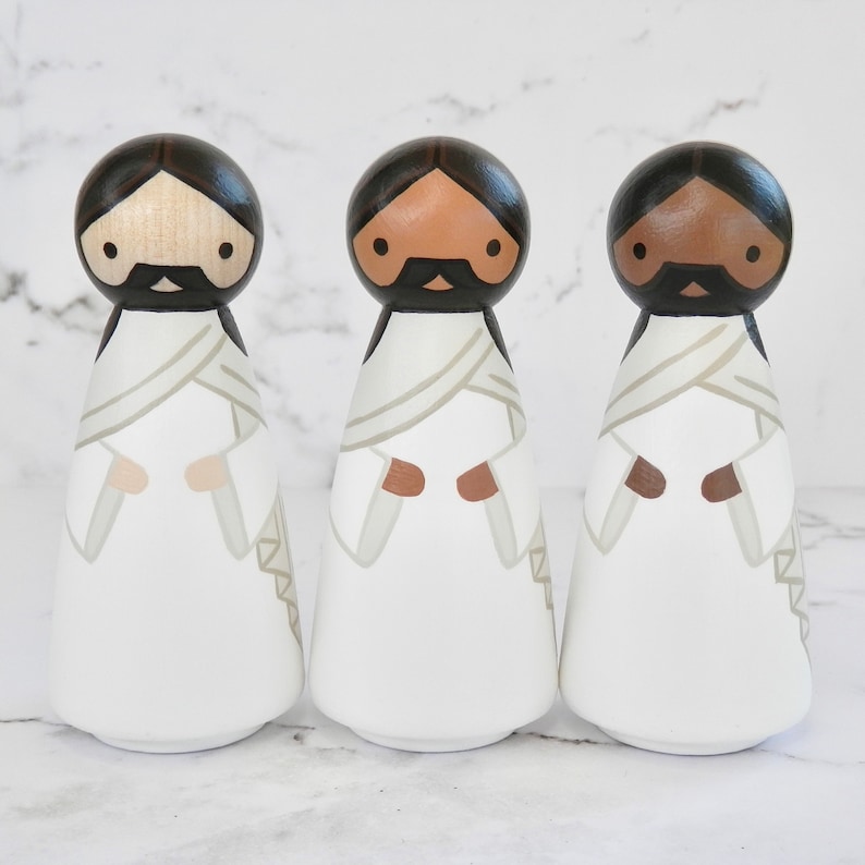 May include: Three wooden peg dolls dressed in white robes with brown, tan, and black painted faces. The dolls have black hair and black mustaches.