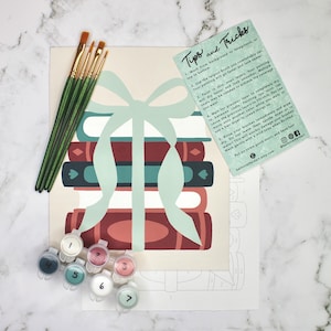 May include: A paint-by-numbers kit featuring a stack of books tied with a ribbon. The kit includes paint pots, paintbrushes, and a guide with instructions. The color palette includes shades of red, teal, and cream. The kit is designed for artistic activities.