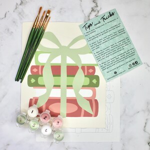 May include: A paint-by-numbers kit featuring a stack of books with a light green ribbon. The kit includes paint pots, paintbrushes, and a guide with tips and tricks. The color palette includes shades of red, green, and pink.