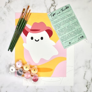 May include: A paint-by-number kit featuring a cartoon ghost wearing a pink cowboy hat. The kit includes paint pots, paintbrushes, and a canvas with a numbered outline. The background is yellow and pink with the text "Tips and Tricks".