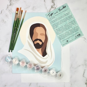May include: A paint-by-numbers kit featuring an illustration of Jesus with a halo, paintbrushes, and numbered paint pots. The kit includes a card with painting tips and tricks. The color palette includes shades of blue, brown, and white.