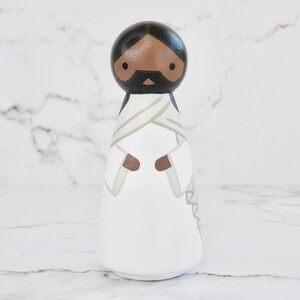 May include: A wooden peg doll painted to resemble Jesus. The doll has a brown skin tone, black hair, and a white robe with a brown sash. The doll's eyes are black, and it has a black beard and mustache.