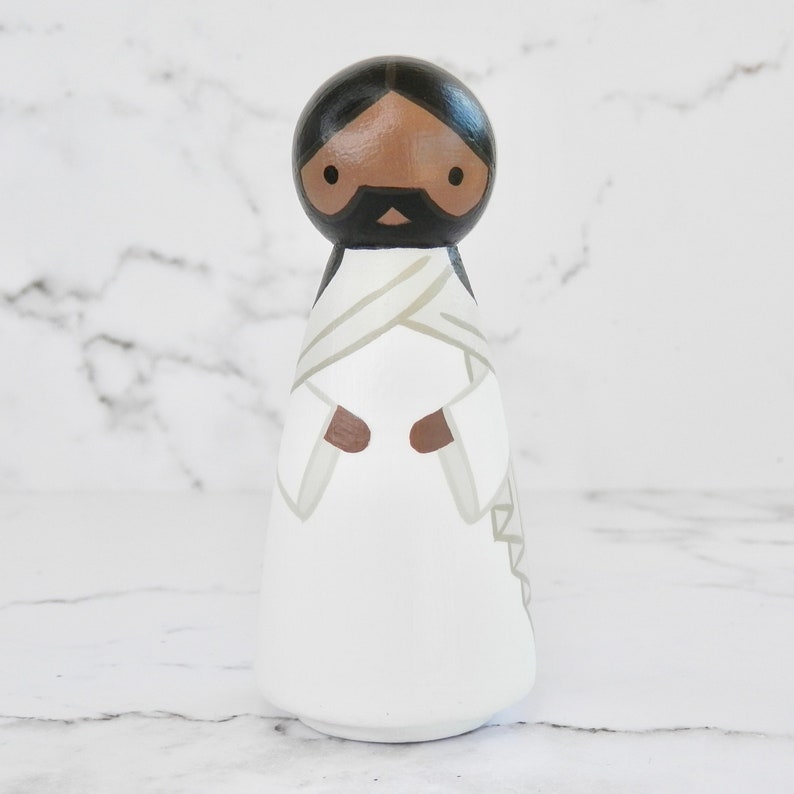 May include: A wooden peg doll painted to resemble Jesus. The doll has a brown skin tone, black hair, and a white robe with a brown sash. The doll's eyes are black, and it has a black beard and mustache.