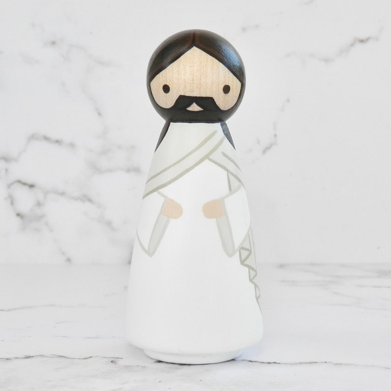May include: A wooden peg doll painted to resemble Jesus. The doll has a white robe with a gray sash, brown hair, and a brown beard.