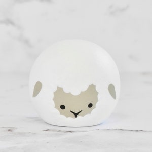 May include: A white ceramic sheep figurine with a gray face and black eyes. The sheep has a simple, rounded shape and a fluffy, white wool texture.