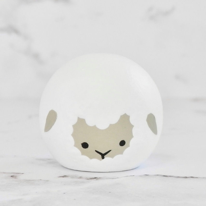 May include: A white ceramic sheep figurine with a gray face and black eyes. The sheep has a simple, rounded shape and a fluffy, white wool texture.