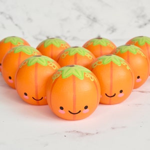 May include: Eight hand-painted wooden pumpkins with smiling faces. Each pumpkin is painted orange with a green stem and a pink blush on the cheeks.