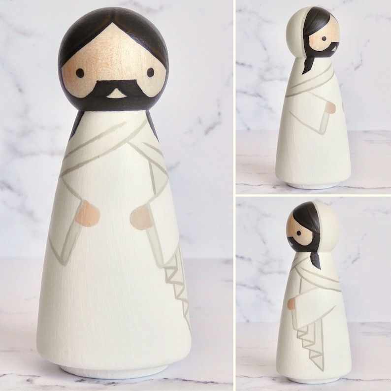 May include: Hand-painted wooden doll with black hair and a white robe. The doll has a simple, stylized face with black eyes and a black beard. The doll is standing on a white background.