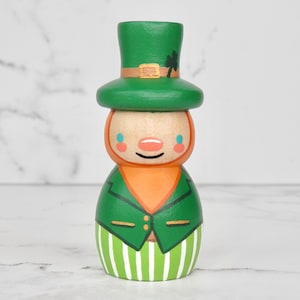 May include: A wooden figurine of a leprechaun wearing a green hat with a gold band and a shamrock. The leprechaun has a green jacket, orange shirt, and green and white striped pants.