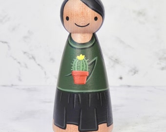 May include: A wooden doll with black hair and a green shirt with a cactus design. The doll is wearing a black skirt and has a painted smile.