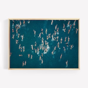 Op de afbeelding: Een luchtfoto van een groep mensen die zwemmen in een turquoise blauwe oceaan. De zwemmers zijn verspreid over de hele afbeelding, wat een dynamische en kleurrijke compositie creëert.
