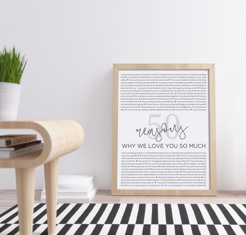 May include: A framed print with a white background and black text. The text reads "50 reasons why we love you so much". The print is on a wooden table with a black and white rug.