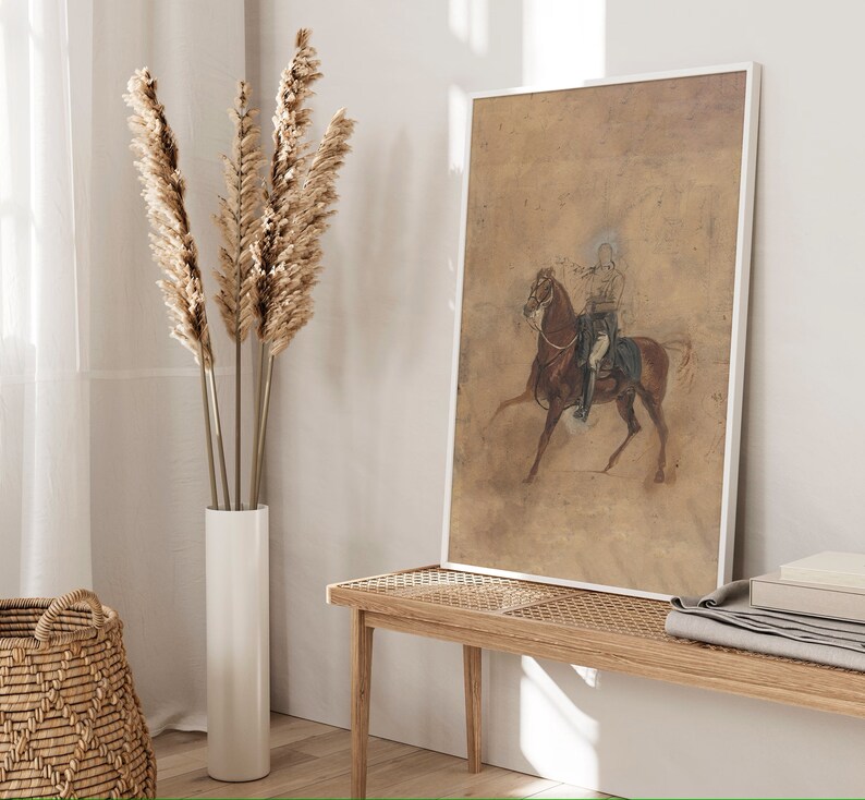 May include: A framed print of a vintage illustration depicting a person riding a horse. The print is on a beige background and features a brown horse with a rider wearing a blue and white uniform. The print is framed in a white frame.