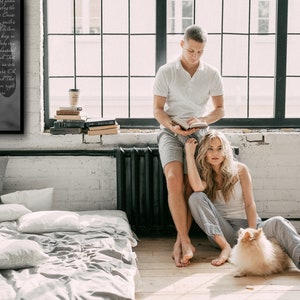 May include: A couple sits on the floor in a loft-style room with a large window. The woman is sitting with her legs crossed and her arms around a small, fluffy dog. The man is standing behind her, leaning against a radiator, and reading a book. There is a bed in the background and a framed print on the wall with the text "Jonathan & Ricardo, November 12, 2021".