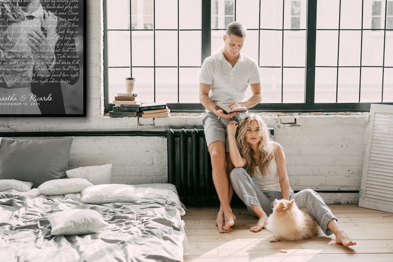 May include: A couple sits on the floor in a loft-style room with a large window. The woman is sitting with her legs crossed and her arms around a small, fluffy dog. The man is standing behind her, leaning against a radiator, and reading a book. There is a bed in the background and a framed print on the wall with the text "Jonathan & Ricardo, November 12, 2021".