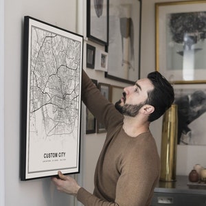 May include: A person hangs a black framed map print on a white wall. The map print features a black and white city map with the text "CUSTOM CITY" and "LOCATION TO YOUR PLACE".