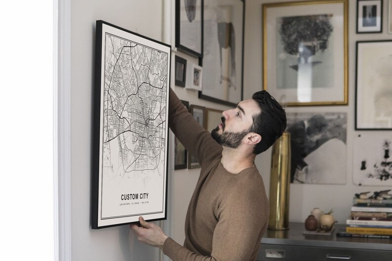 May include: A person hangs a black framed map print on a white wall. The map print features a black and white city map with the text "CUSTOM CITY" and "LOCATION TO YOUR PLACE".