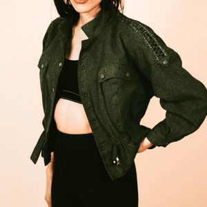 Könnte beinhalten: Eine dunkelgrüne, langärmelige Jacke mit Knopfleiste und Kordelzug in der Taille. Die Jacke hat einen strukturierten Stoff und eine kleine Tasche auf der linken Seite.