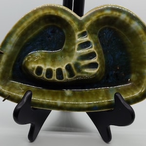 May include: Green and blue ceramic ashtray shaped like a foot. The ashtray has a glazed finish and is decorated with a foot print.